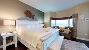 A bedroom with a large bed, a nightstand, a chair, and a view of the sea.