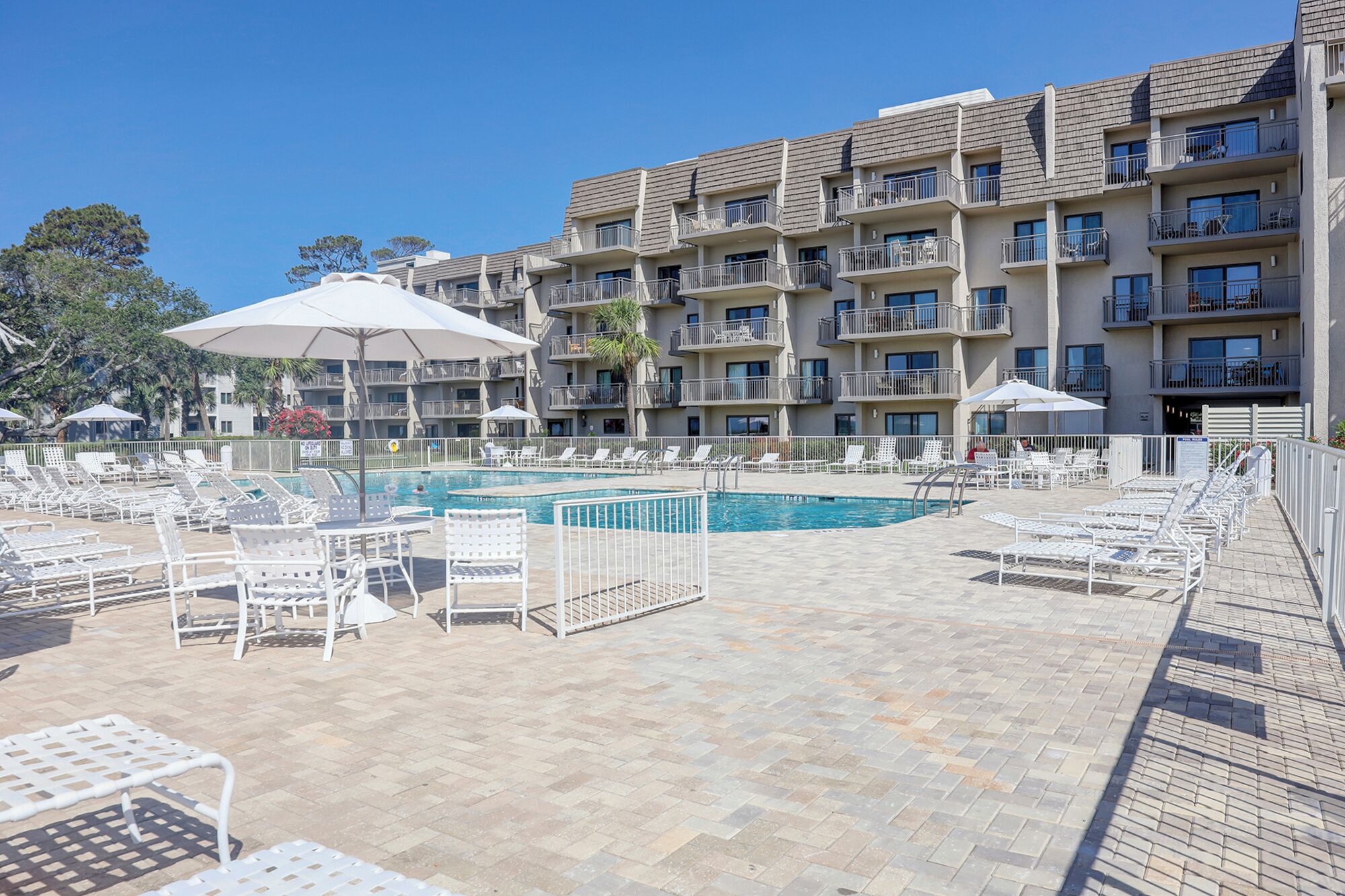 A hotel pool area with multiple lounge chairs and umbrellas, surrounded by a multi-story building with balconies.
