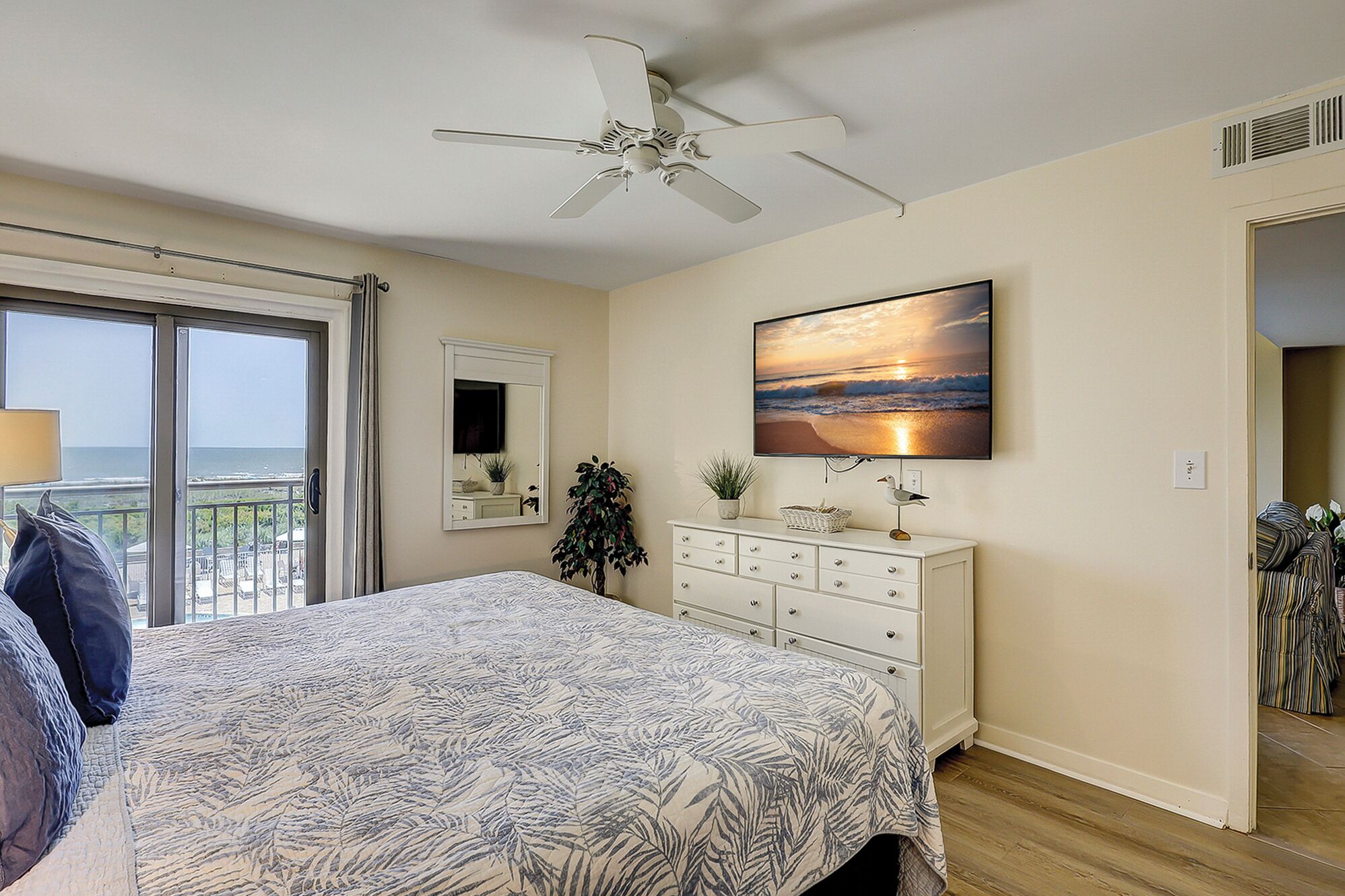 Une chambre avec un lit, un ventilateur de plafond, une télévision, une commode et une vue sur l’océan, accessible par une porte coulissante.