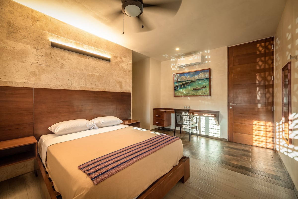 A bedroom with a large bed, wooden headboard, bedside tables, a desk with a chair, a painting on the wall, and a sliding door.