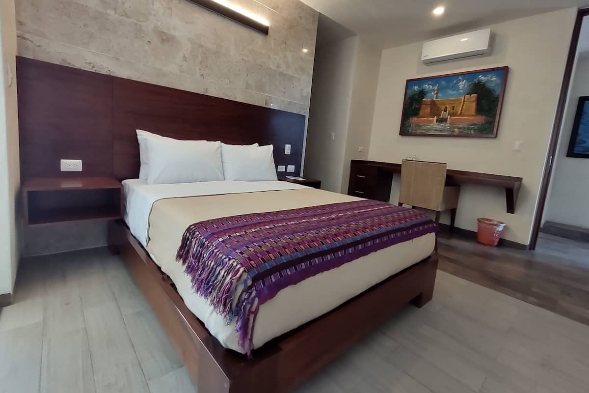 A bedroom with a large bed, a wooden headboard, a nightstand, a desk, and an air conditioning unit.