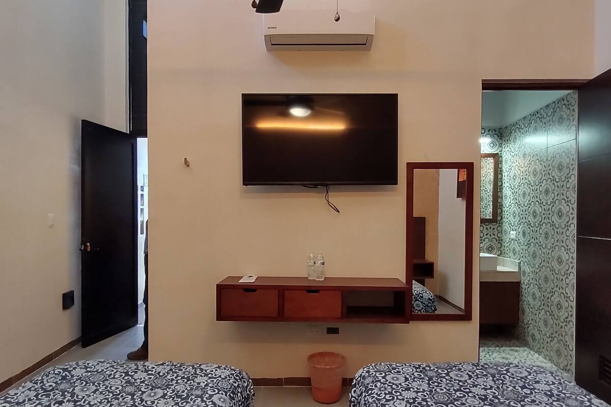 A hotel room with two beds, a ceiling fan, a TV, a minibar, and a bathroom visible through an open door.