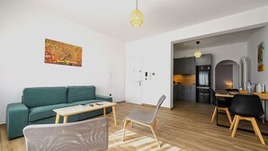 A modern living room with a green sofa, a wooden coffee table, and a grey chair. A kitchen is visible in the background with a dining table and chairs. - Alexander Apartments Airport (Spata-Artemida)