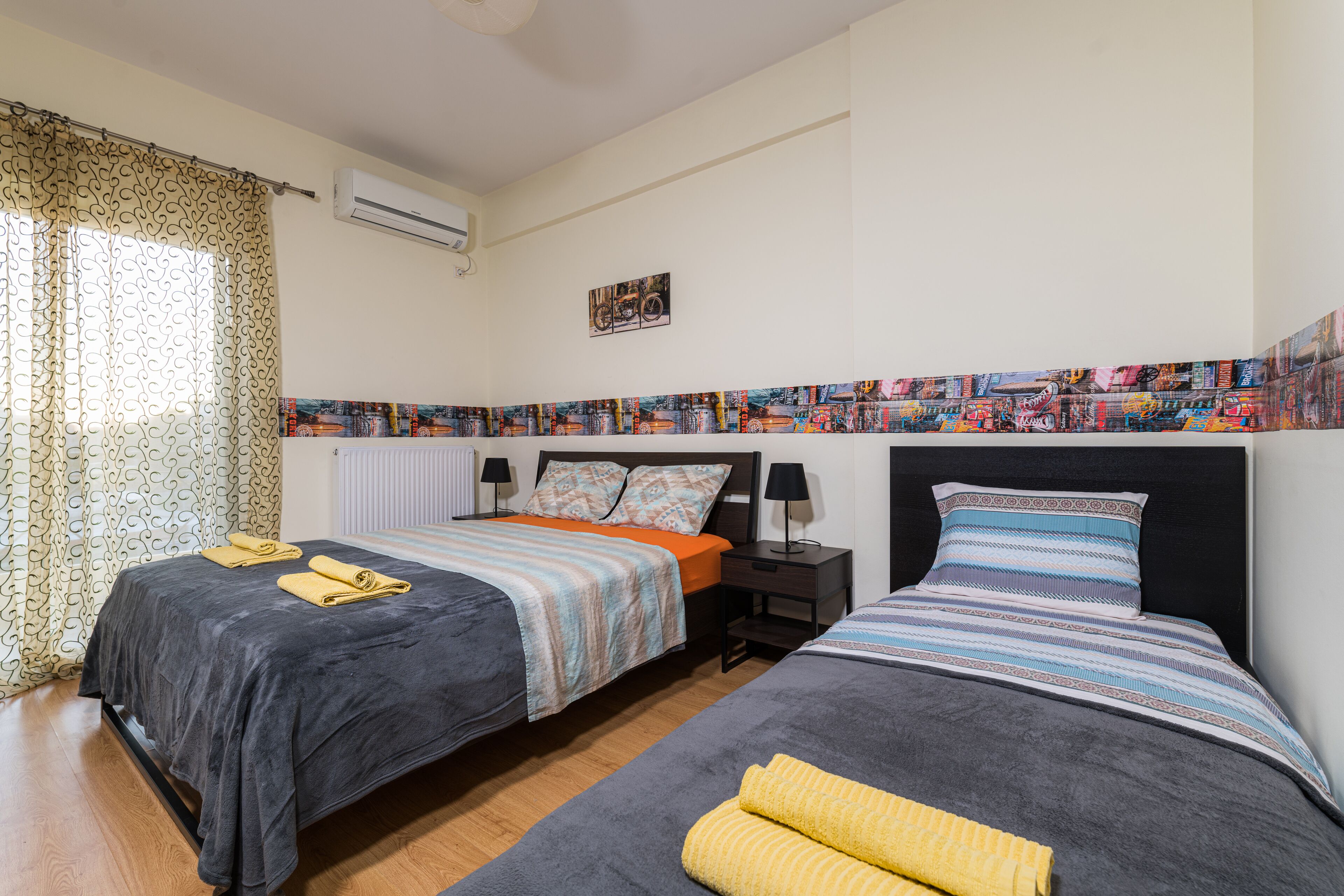 A hotel room with two beds, a patterned headboard, a radiator, and a window with curtains.