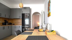 A modern kitchen with grey cabinets, a wooden dining table with black placemats, and a hanging light fixture. - Alexander Apartments Airport (Spata-Artemida)
