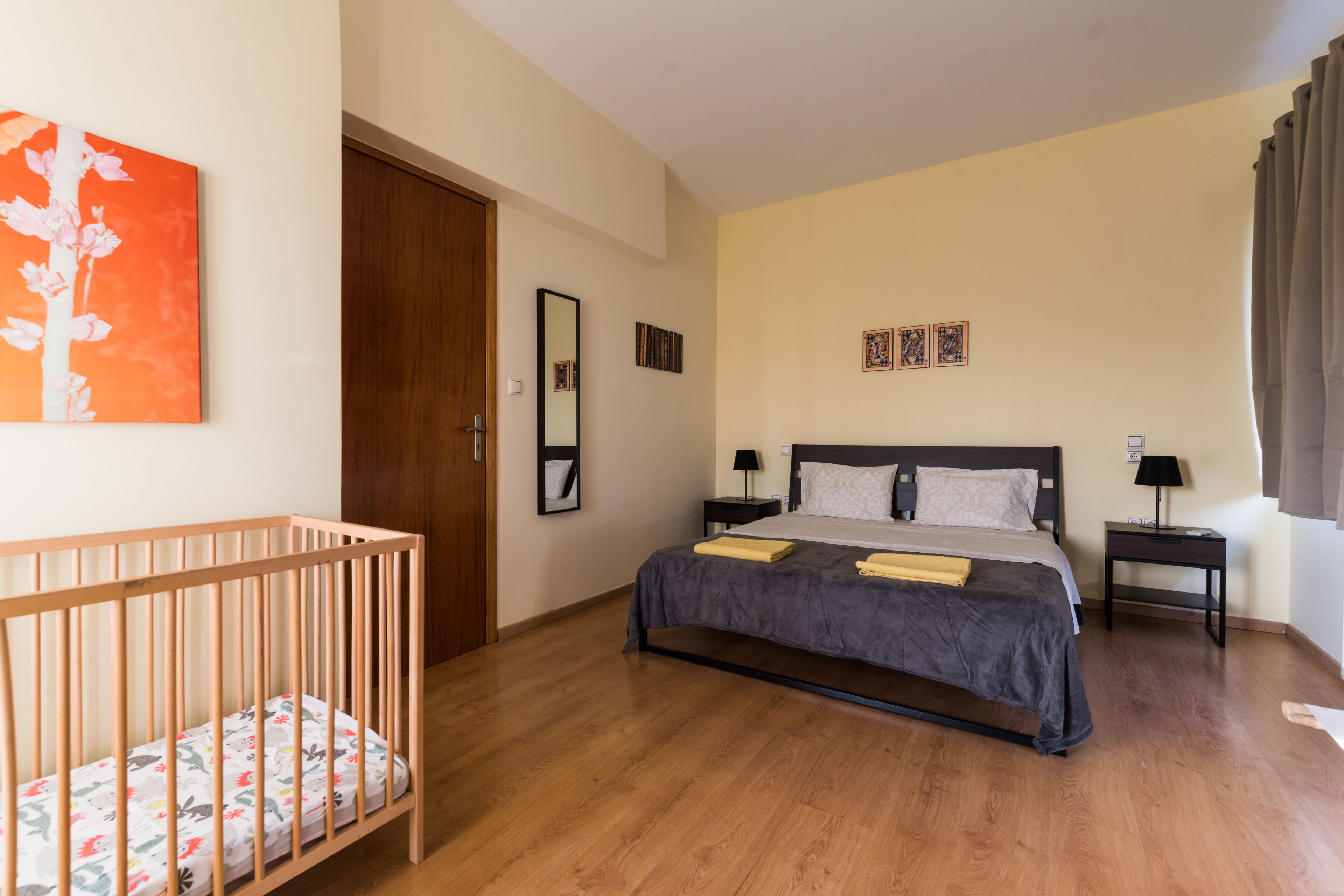 Une chambre avec un berceau en bois, un lit recouvert d’une couverture grise, deux tables de chevet avec des lampes et un grand miroir au mur.