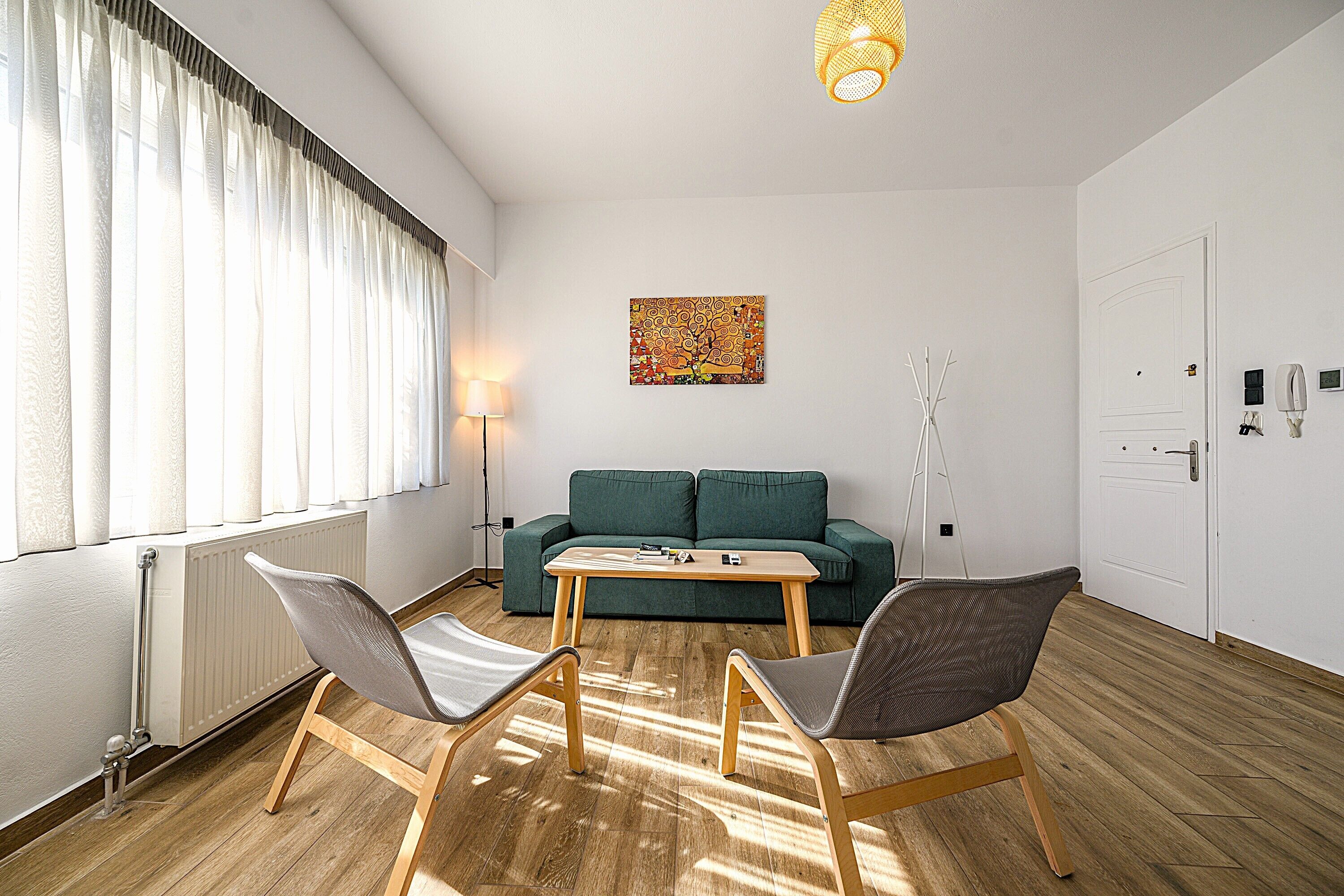 A room with a wooden floor, a green sofa, two chairs, a table, a lamp, a painting, and a door.