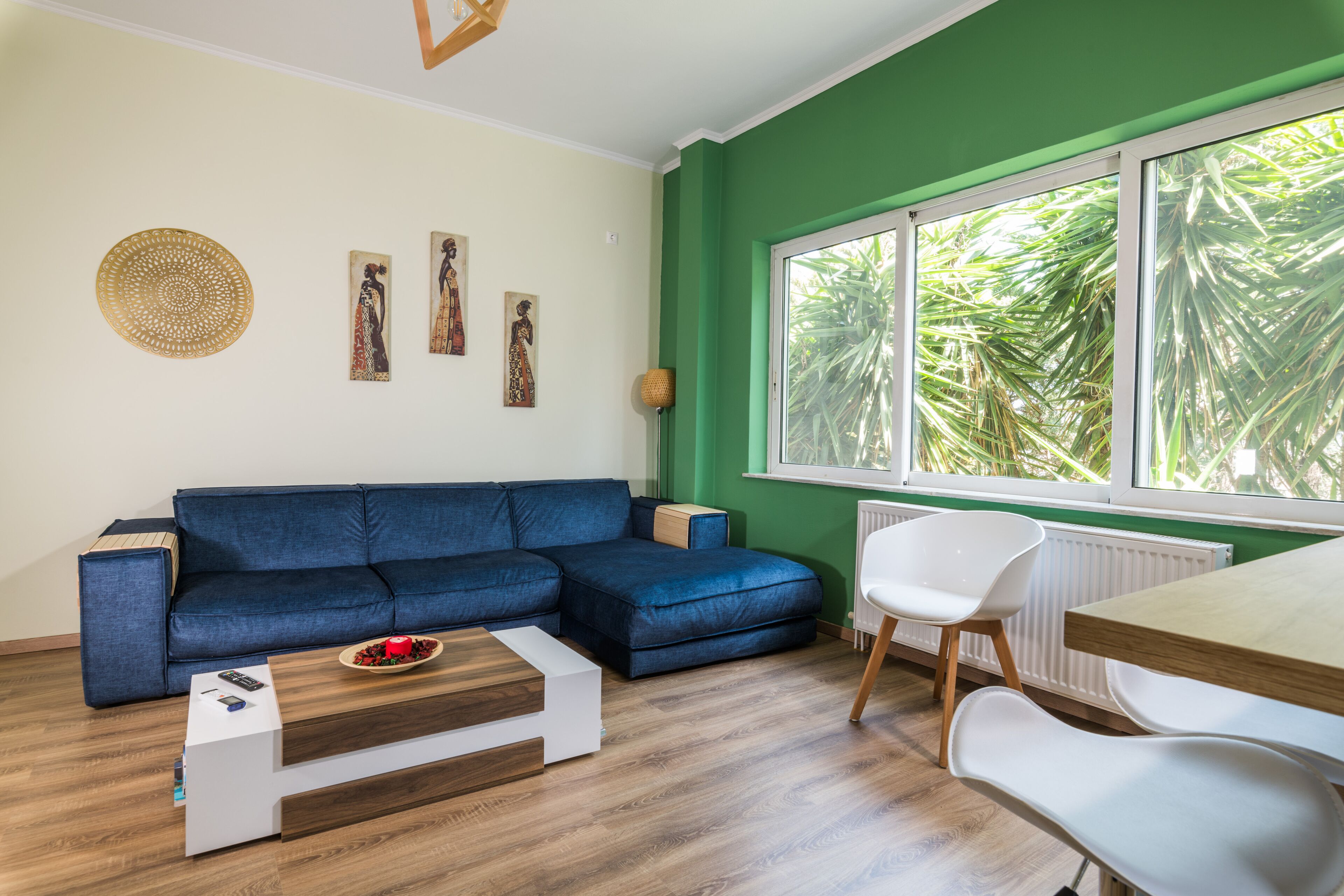 A modern living room with a blue sofa, a wooden coffee table, and a white chair. There are three wall-mounted artworks and a large window with a view of palm trees.