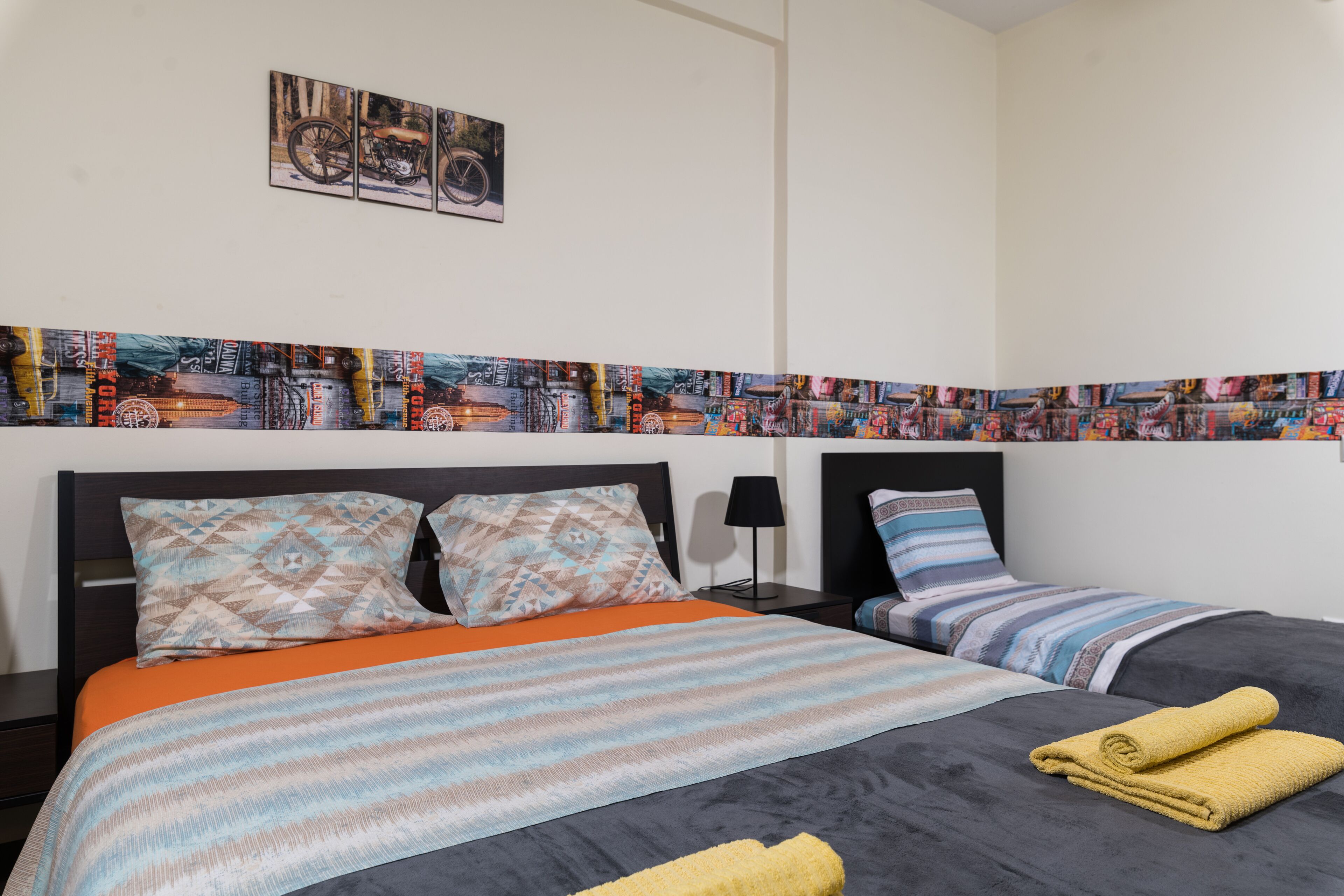 A hotel room with two beds, a patterned headboard, a cityscape artwork, and a nightstand with a lamp.