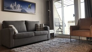 A living room with a grey sofa, a brown armchair, a large window with curtains, and a framed picture of a snowboarder on a mountain. - Mt-Orford ski 8.5Km. Walk to old Magog! Lake view w/pools, hot tub, sauna! Quiet (Magog)