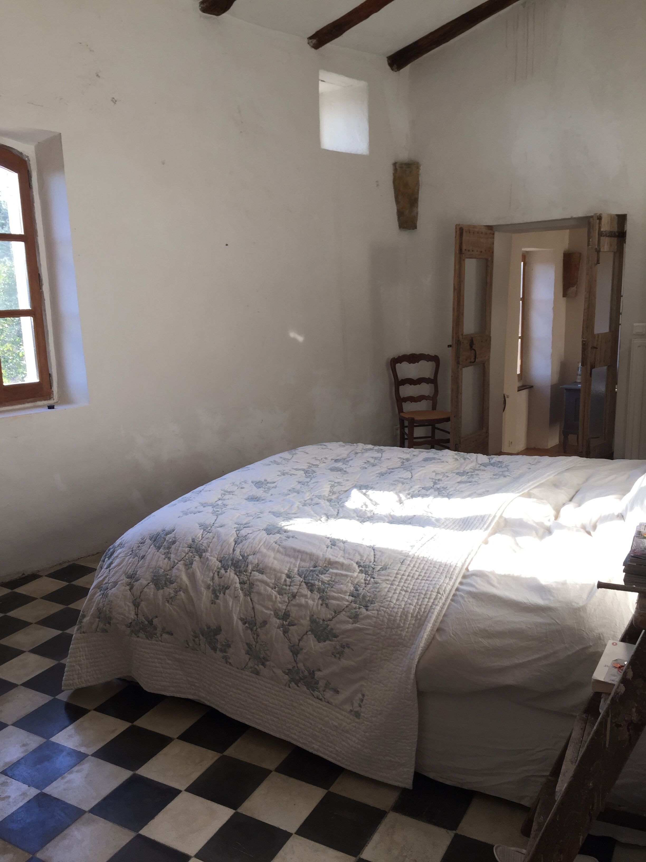 A room with a checkered floor, a bed with a floral bedspread, a wooden chair, and a door with a mirror.