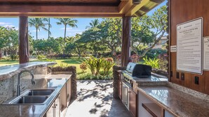 Eine Außenküche mit Stein Arbeitsplatte, Spüle und Grill, mit Blick auf einen angelegten Garten mit Palmen und einem Pool.