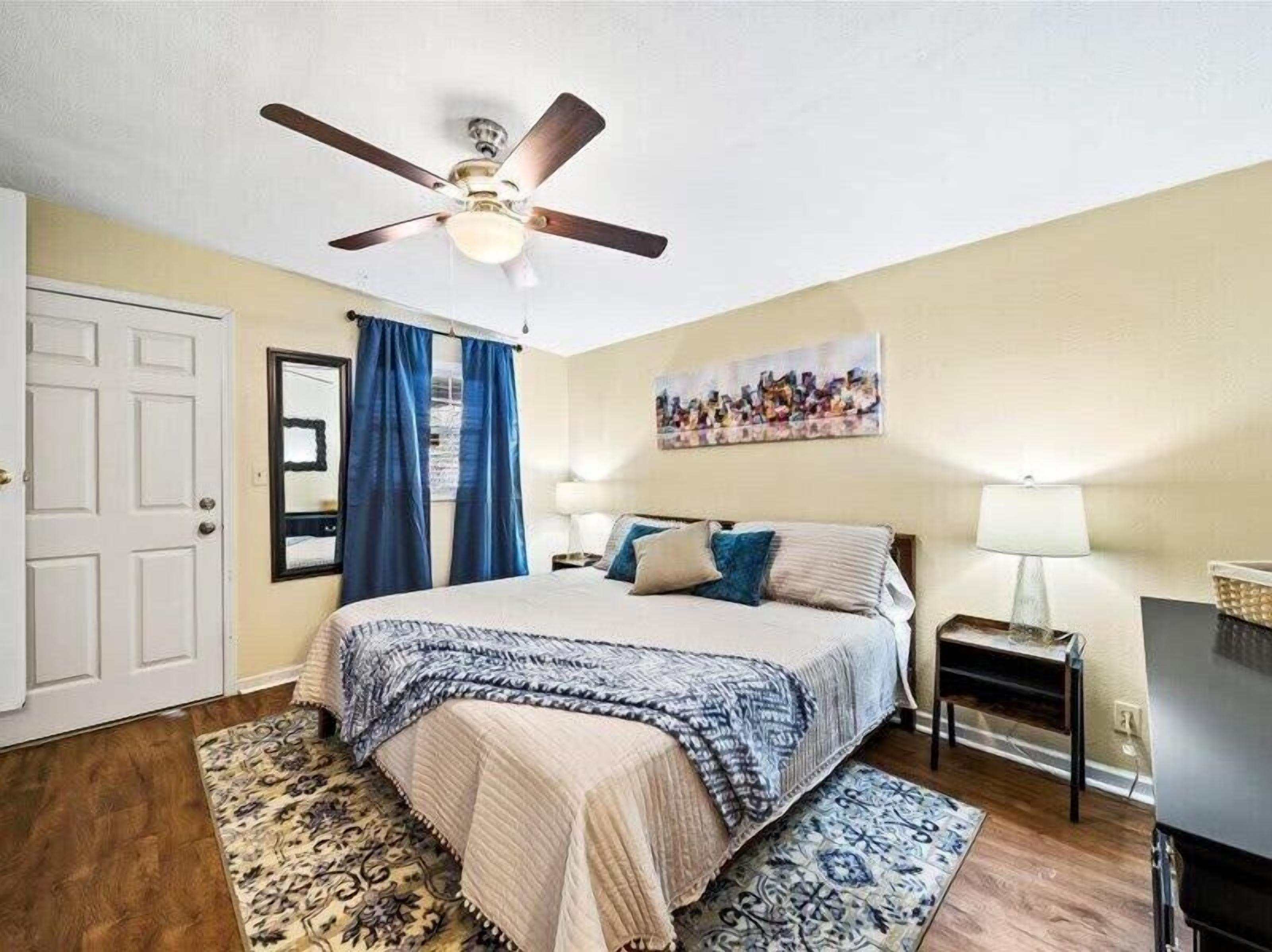 Une chambre à coucher avec un lit, un ventilateur de plafond, une table de chevet, une lampe, un tableau et une porte.