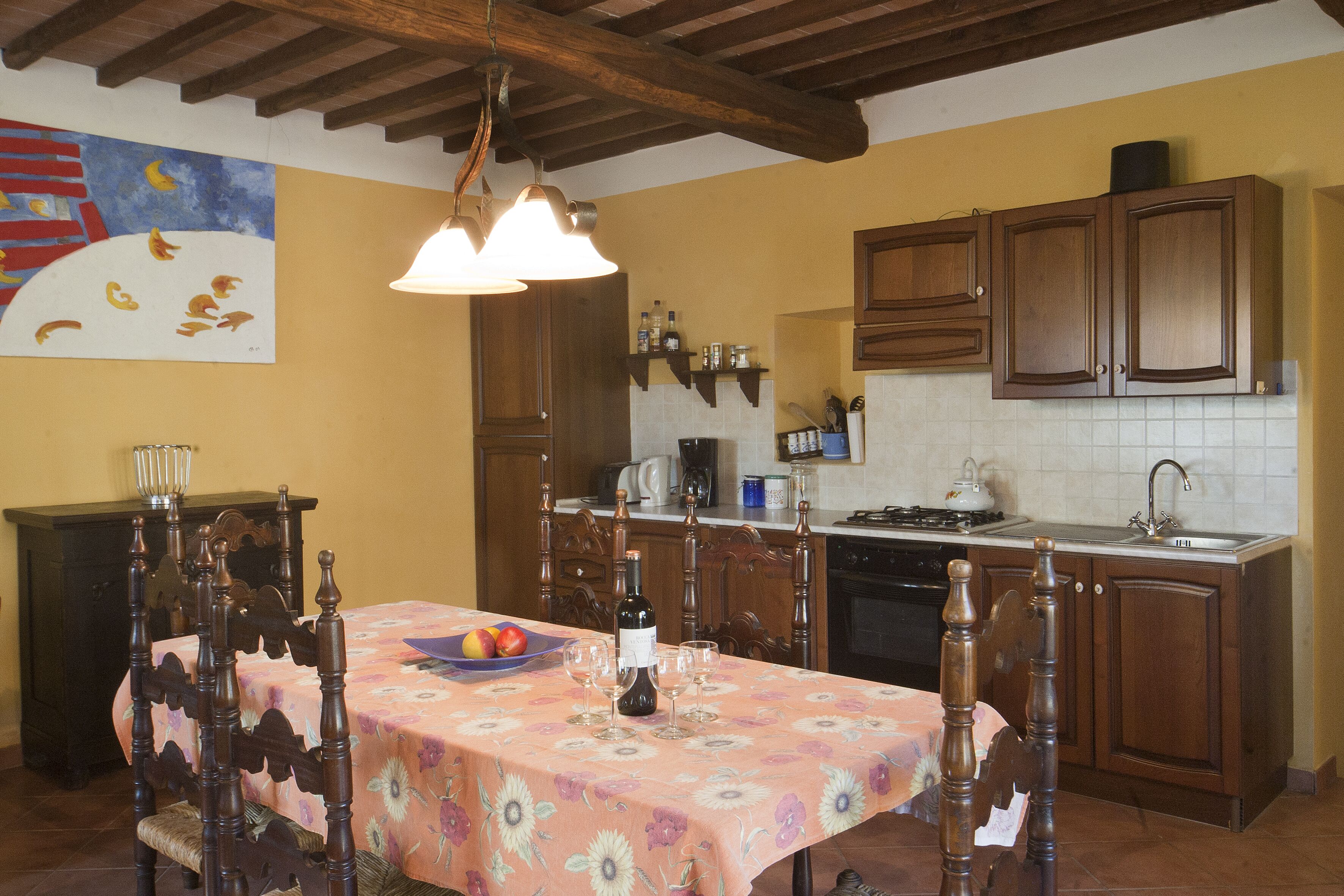 A dining area with a table set for six, a painting on the wall, wooden cabinets, and a kitchen in the background.