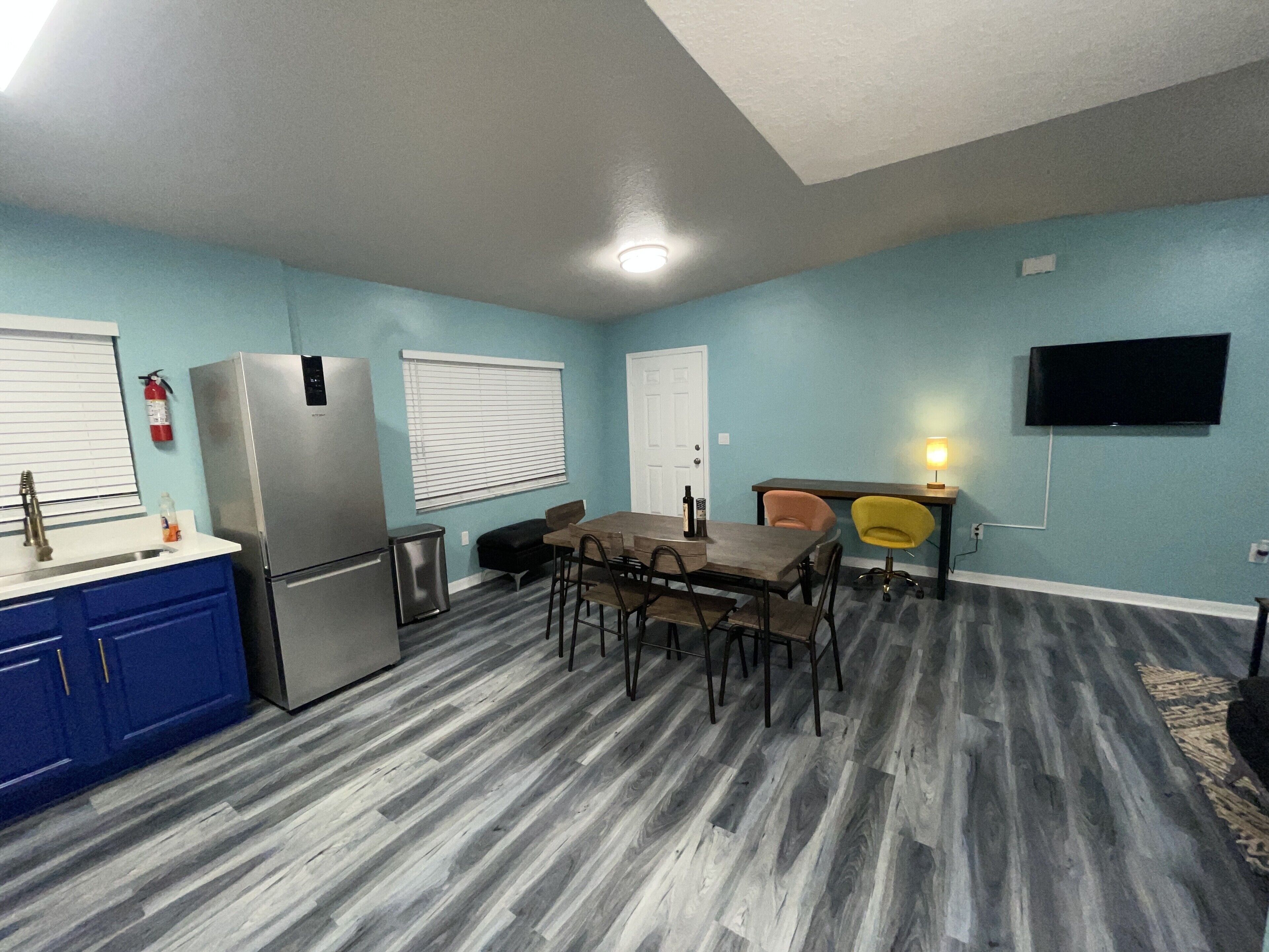 A compact kitchen with a refrigerator, sink, and a small trash bin. There is a dining area with a table and chairs, and a television mounted on the wall.