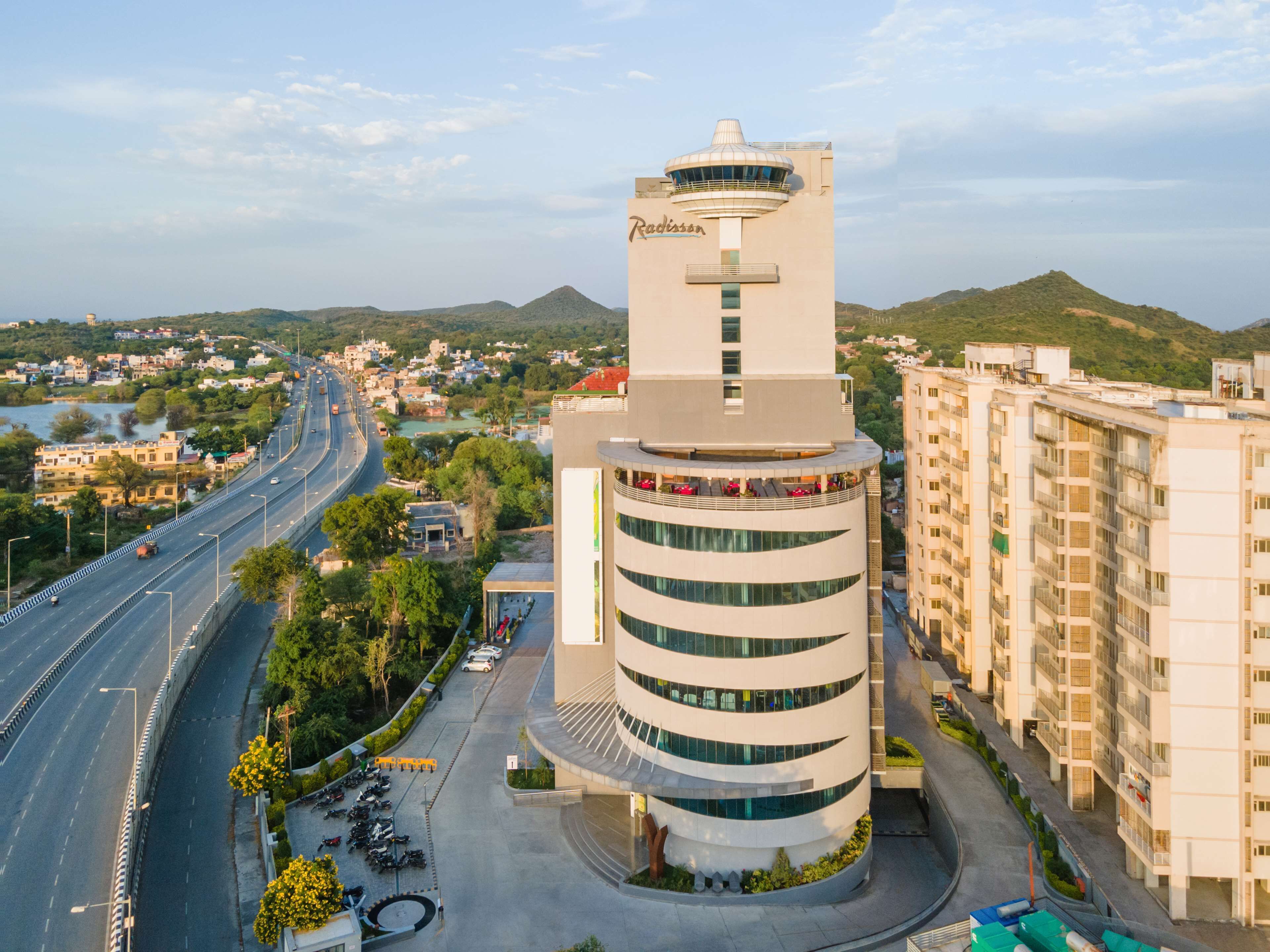 Un hotel moderno con una torre central, un área de estacionamiento y vistas a una autopista y el paisaje circundante.
