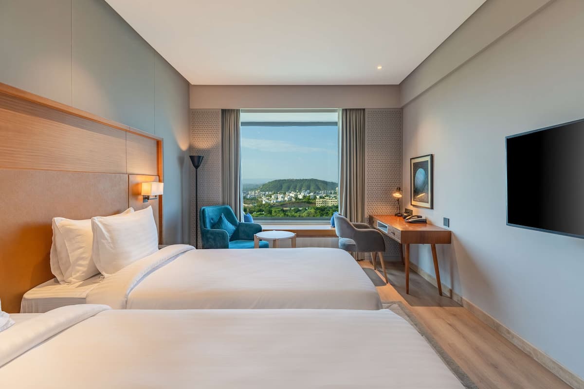 A hotel room with two beds, a desk, a chair, a TV, and a large window with a city view.