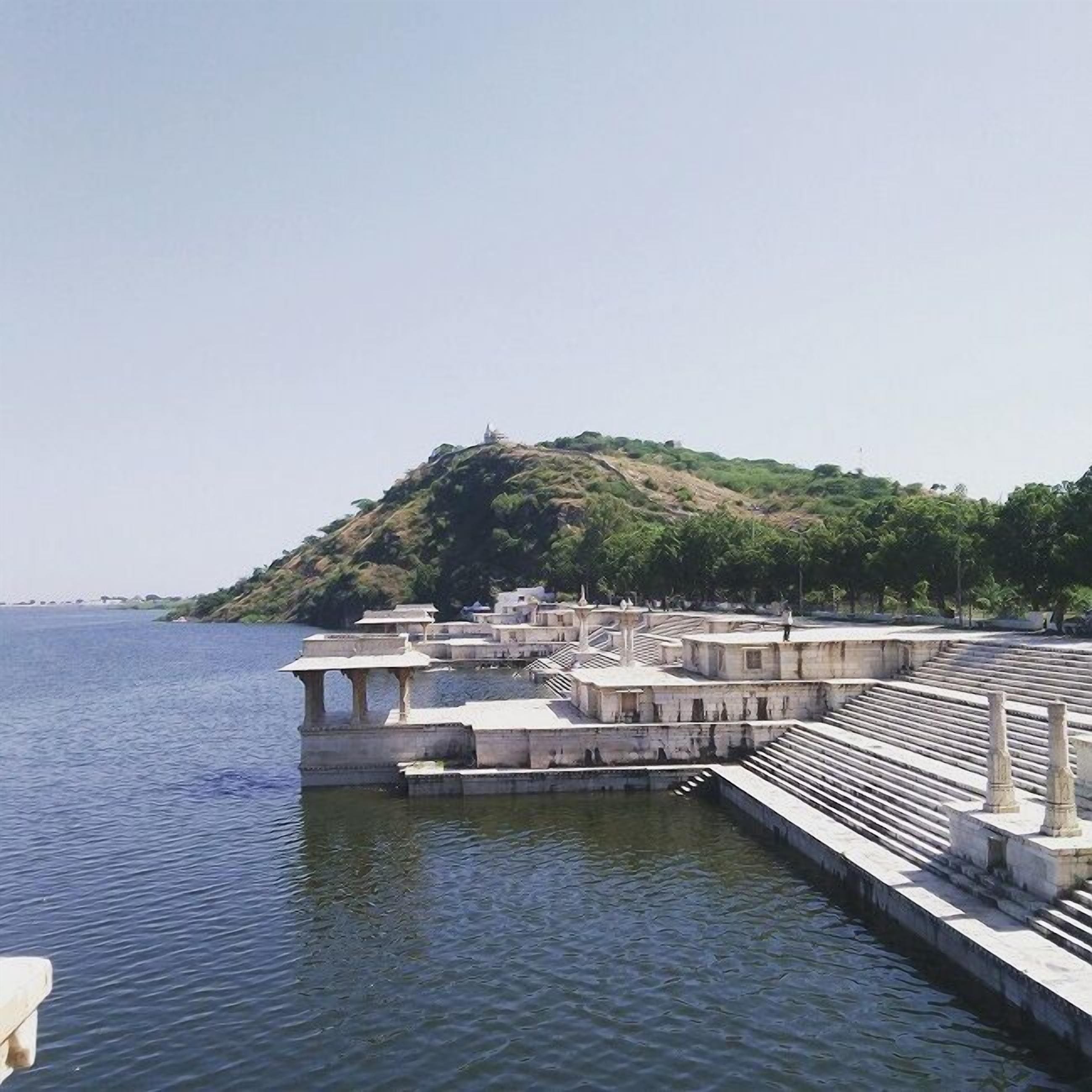 Ein Ufergebiet mit einer weitläufigen Freifläche, Treppen, die zum Wasser führen, und einem Hügel mit einer Statue im Hintergrund.