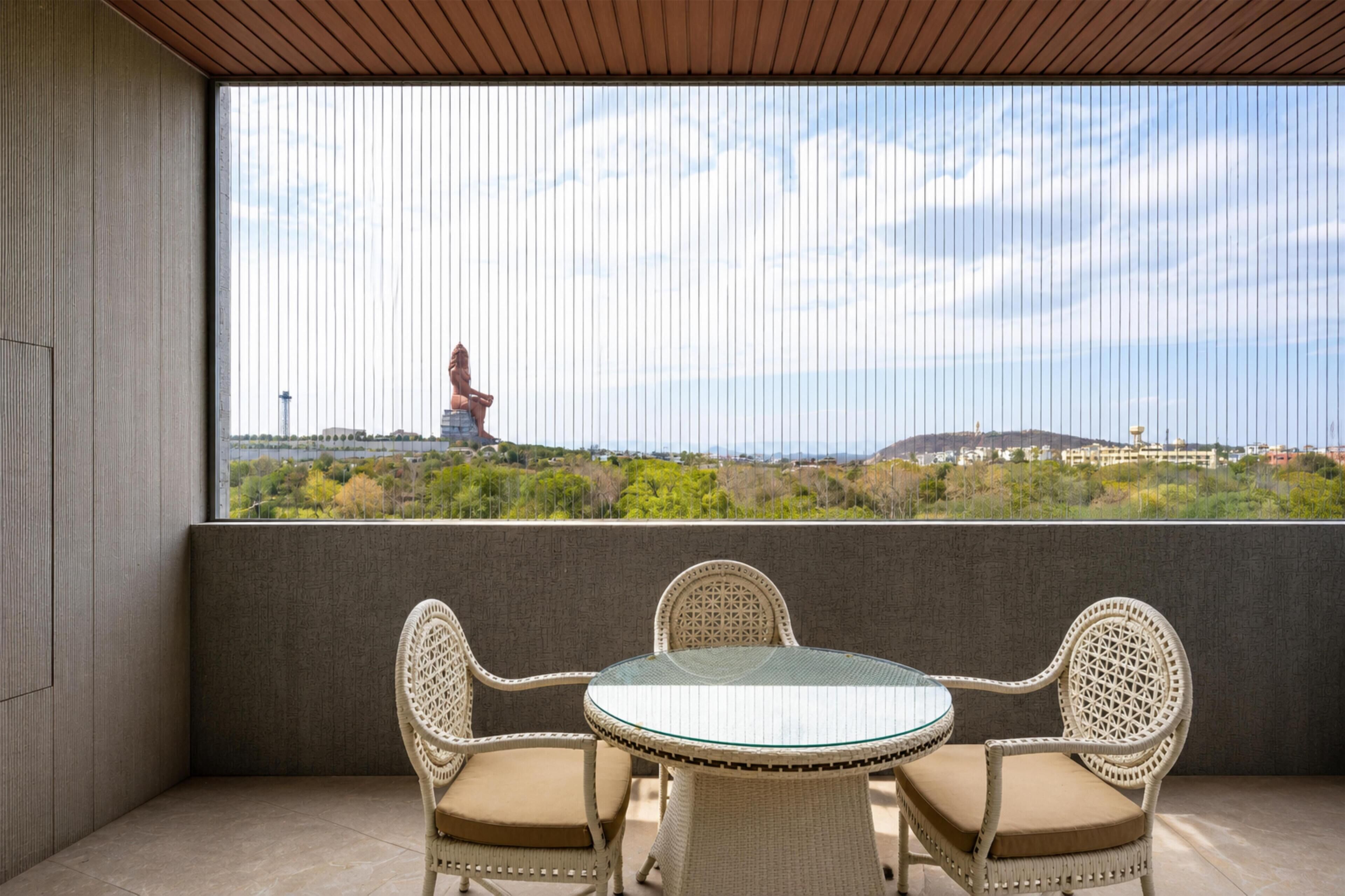 A balcony with wicker chairs and a glass table, overlooking a scenic view with a statue in the distance.