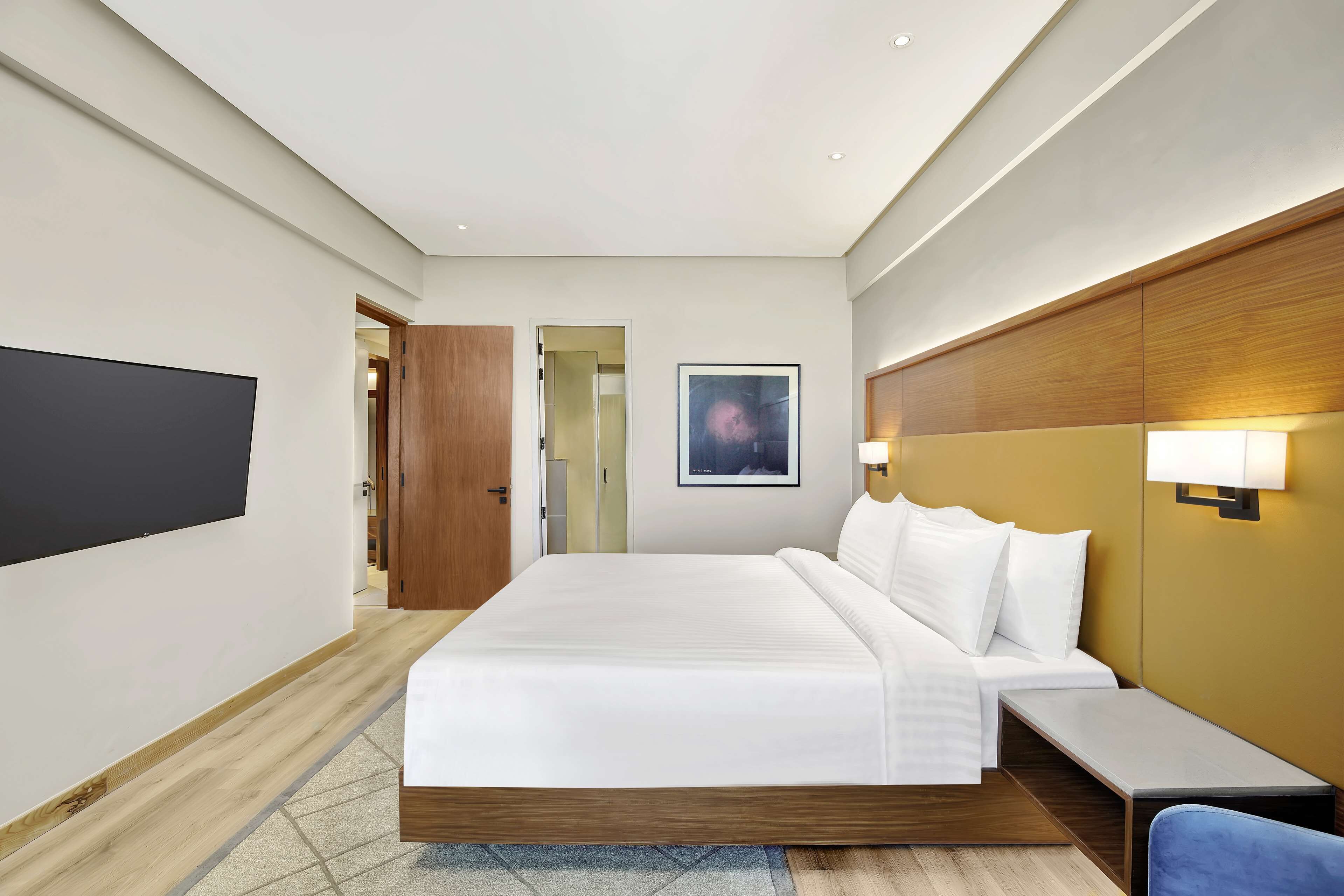 Ein modernes Hotelzimmer mit einem großen Bett, einem Nachttisch, einem an der Wand montierten Fernseher und einer Tür, die in einen weiteren Raum führt.