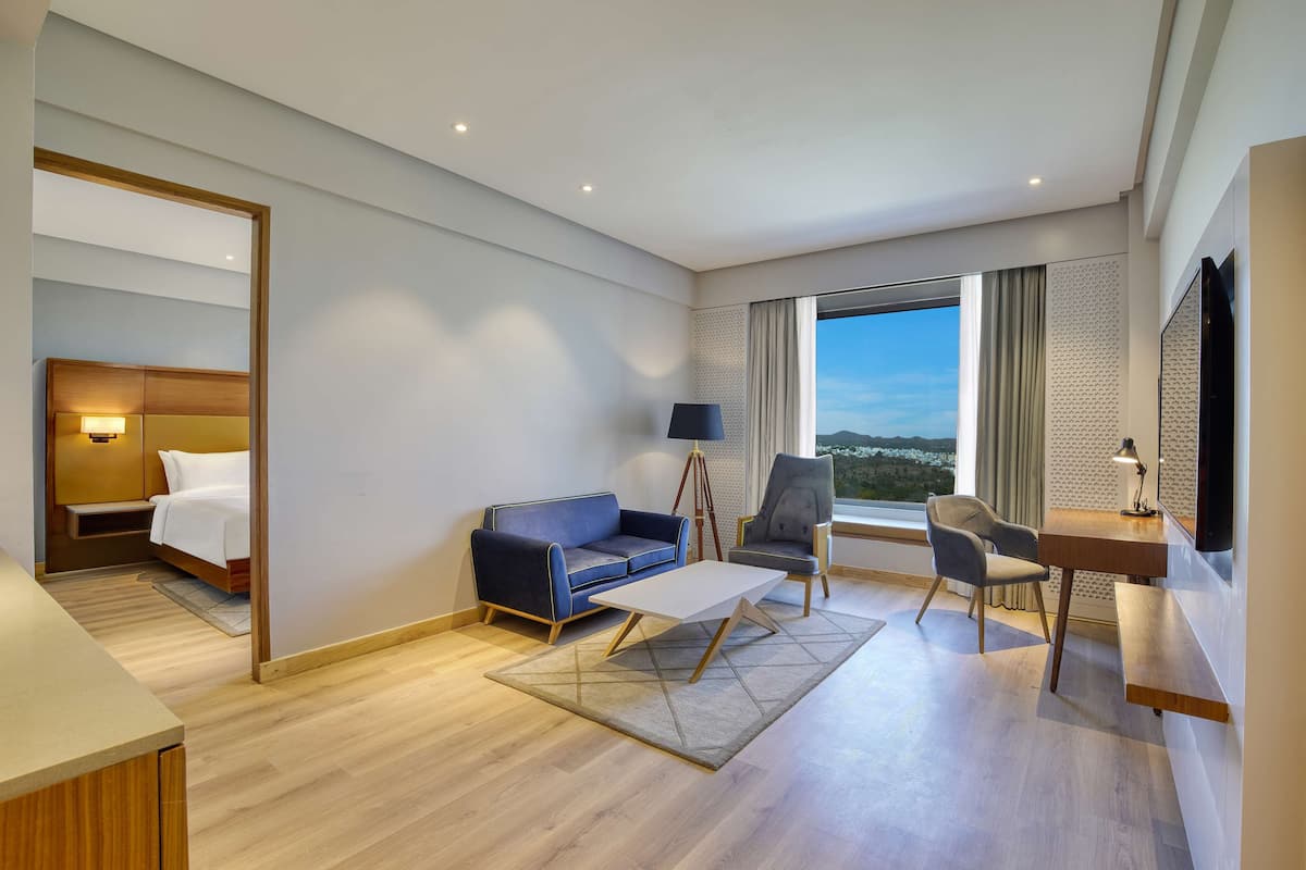 A modern hotel room with a bed, a blue sofa, a desk with a lamp, a chair, and a large window offering a view.