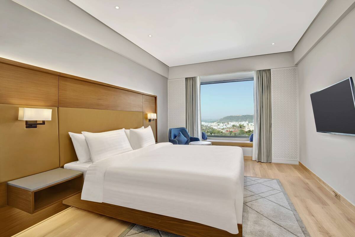 A modern hotel room with a large bed, bedside tables, a TV, and a view of a cityscape.