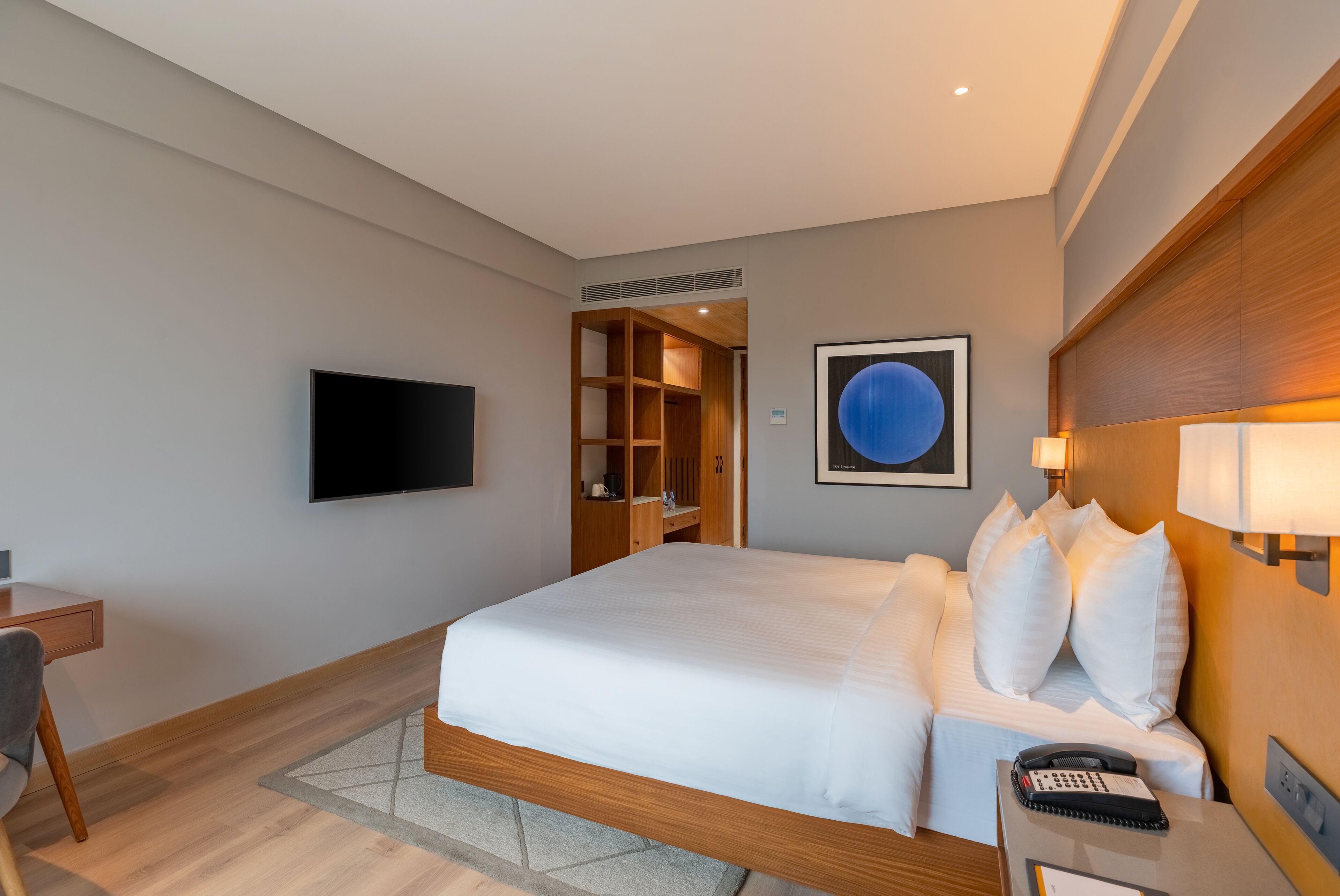 A modern hotel room with a large bed, a desk, a flat-screen TV, a wooden headboard, and a wall-mounted artwork.