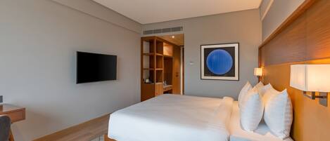 A modern hotel room with a large bed, a desk, a flat-screen TV, a wooden headboard, and a wall-mounted artwork.