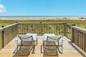 Une terrasse en bois avec deux chaises longues blanches et une table, donnant sur une plage. On aperçoit une clôture et des véhicules au loin.