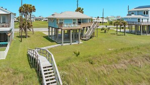 A beachfront house with a wooden deck, a staircase leading to a balcony, and a grassy area. - Updated Beachfront Getaway w/ 750 sq ft Deck, Private Walkover & Great Sunrises! (Galveston)