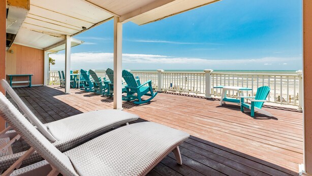 Een houten terras met lounge fauteuils en een tafel, met uitzicht op een strand.