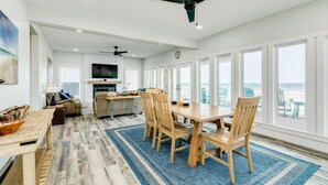 Een ruime woonkamer met een grote eettafel, houten stoelen en een blauw vloerkleed. Er zijn plafondventilatoren, een televisie en uitzicht op de oceaan door grote ramen.