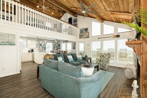 A spacious living area with a high ceiling, wooden beams, and a balcony with a railing. - Fresh Updates! Beachfront Escape w/ 2 Decks, Open Living Area - Steps from Beach (Galveston)