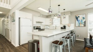 A modern kitchen with white cabinetry, a central island with bar stools, stainless steel appliances, pendant lighting, and a large abstract painting on the wall. - Fresh Updates! Beachfront Escape w/ 2 Decks, Open Living Area - Steps from Beach (Galveston)