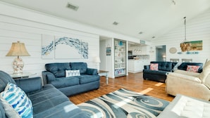 A spacious living room with a blue sofa, a beige armchair, and a patterned rug. A kitchen area is visible in the background with white cabinets and a dining table. - Budget & Pet Friendly Beachfront Home - Great for Couples or Small Families (Galveston)
