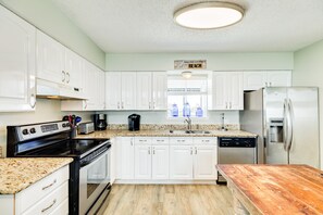 A kitchen with white cabinets, a granite countertop, stainless steel appliances, and a wooden table. - Private Beachfront Home - HOT TUB w/ Gulf Views, Steps from Beach & Dog Friendly (Galveston)