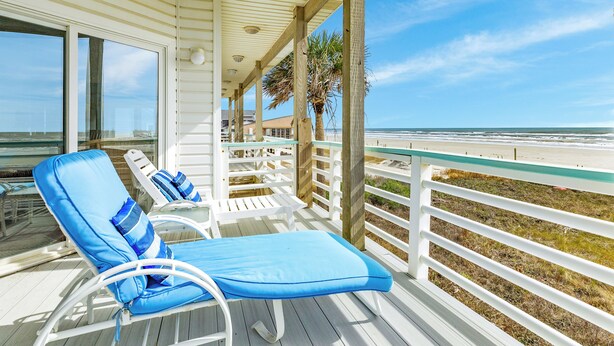 Une terrasse couverte avec deux chaises longues bleues et une porte vitrée donnant accès à la plage.
