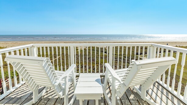 Eine Holzterrasse mit weißen Geländern und zwei weißen Adirondack-Stühlen, die auf einen Strand ausgerichtet sind.
