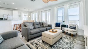 Uma sala de estar moderna com um sofá cinzento, um banquinho e poltronas, uma área de jantar com uma mesa de madeira e grandes janelas com vista.