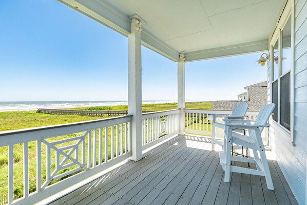 A covered porch with white railings, a view of the beach, and two white Adirondack chairs.