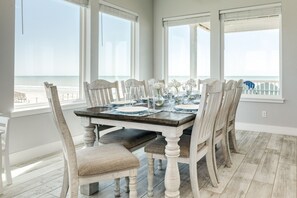 Una sala da pranzo con vista sulla spiaggia, con un tavolo in legno apparecchiato per sei persone, sei sedie imbottite e ampie finestre.