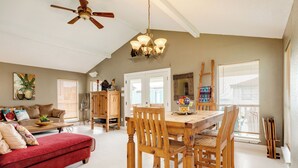 A spacious living room with a red sofa, wooden dining table, and chairs. There is a ceiling fan, a chandelier, and a large window with blinds. - Single Level Beachside Home, Great for Families & Dog Friendly - Walk to Beach (Galveston)