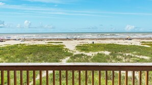 Blick auf den Strand von einer Holzterrasse, mit Dünen und Strandliegen in der Ferne.