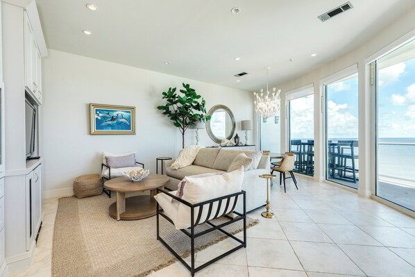 A spacious living room with a large window, a sofa, a coffee table, and a dining area. - 6th Floor Beachfront Condo - Elevator, Private Balcony, Resort Amenities w/ POOL (Galveston)