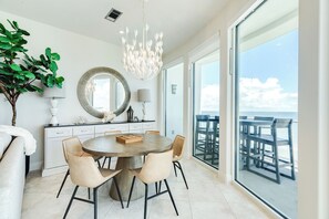 A modern dining area with a round table, beige chairs, a chandelier, a large mirror, and a view of the ocean through large windows. - 6th Floor Beachfront Condo - Elevator, Private Balcony, Resort Amenities w/ POOL (Galveston)