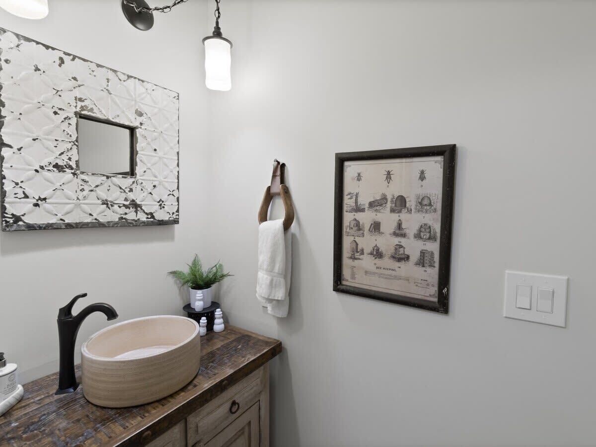 Une salle de bain avec un lavabo rond, un miroir orné d’un motif géométrique, un tableau encadré représentant des bâtiments, une serviette suspendue à un crochet et une plante en pot.