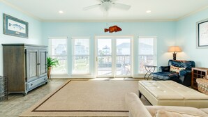 A spacious living room with a large window, a ceiling fan, a fish decoration, and a leather sofa. - Cozy Beachside Home - Pet Friendly, Fenced Yard, Covered Deck, 0.5 blk to Beach (Galveston)