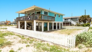 Uma casa de dois andares com varanda, cerca branca de piquete e uma praia arenosa ao fundo.