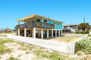 Une maison à deux étages avec un balcon, une clôture blanche en bois et, au loin, une plage de sable.