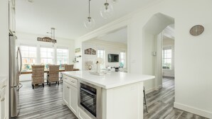A modern kitchen with white cabinetry, a built-in oven, and a large island. There are wicker chairs and a dining table in the adjacent dining area. - Beachside Home w/ Gulf Views Built 2019, 3 Decks, Pointe West Resort Amenities (Galveston)